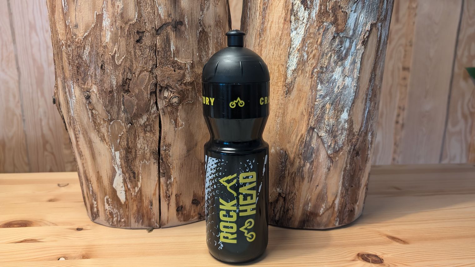 Auf einem Holztisch vor zwei Baumstämmen steht eine schwarze Trinkflasche mit gelber Schrift.