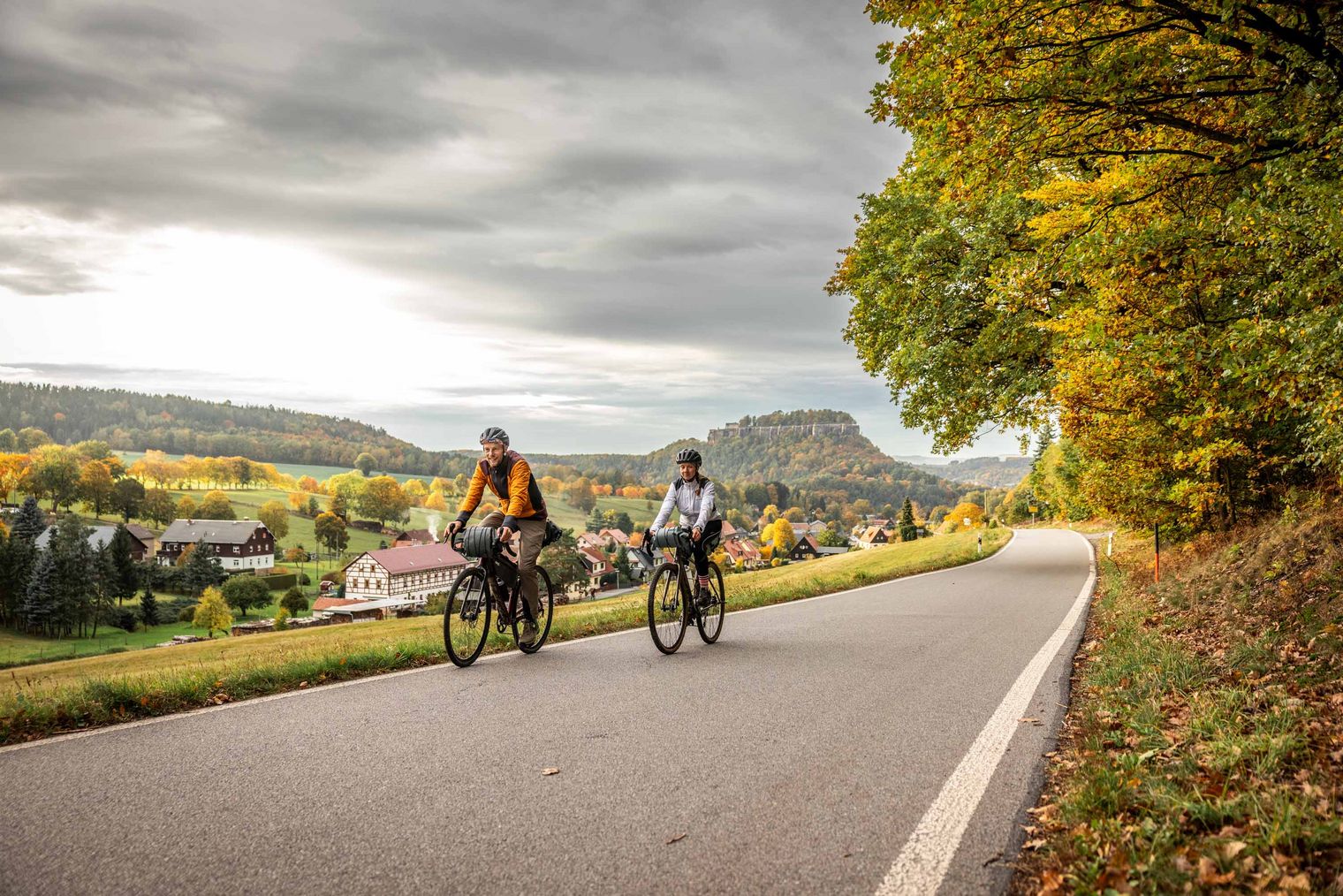 Zwei Radfahrer fahren auf einer Landstraße, umgeben von herbstlichen Bäumen und Hügeln.