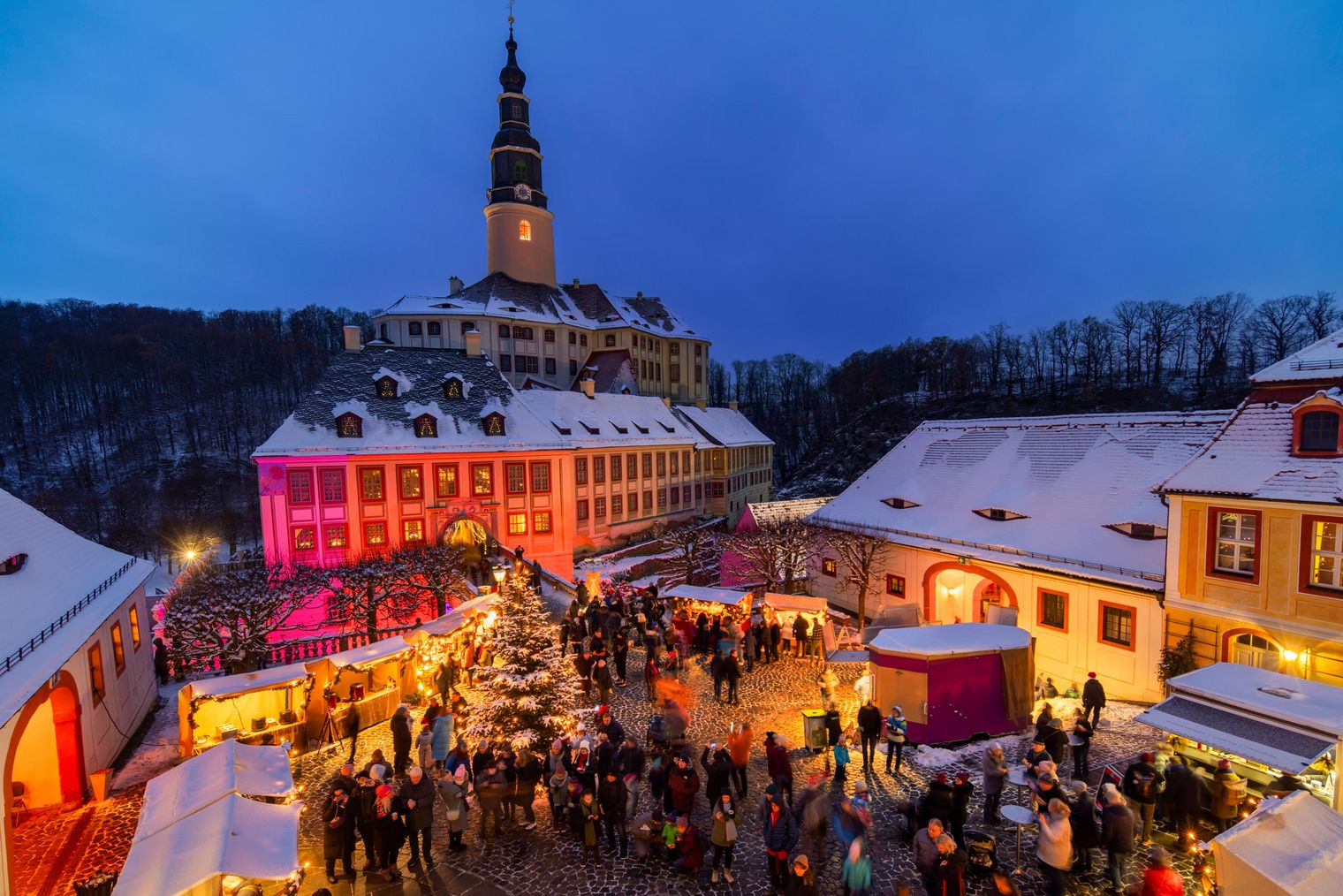 Beleuchteter Weihnachtsmarkt mit vielen Menschen und festlich dekorierten Ständen, umgeben von Schnee.