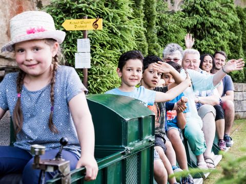 Miniatureisenbahn mit Kindern fährt durch eine grüne Landschaft unter blauem Himmel.