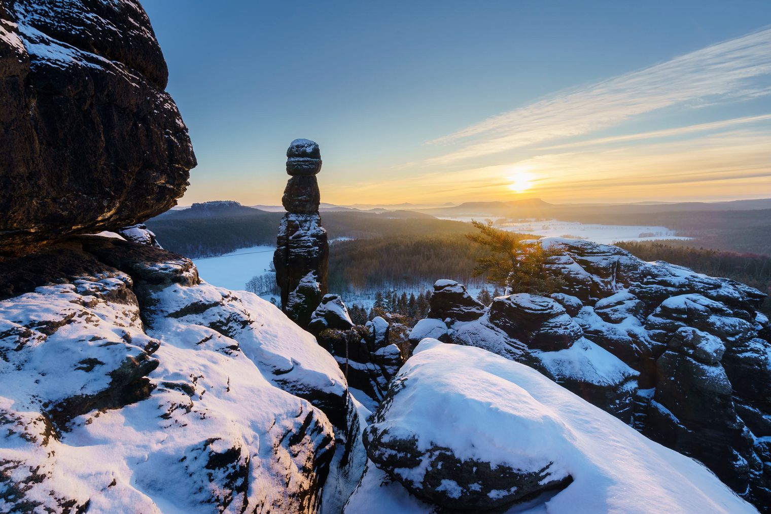 Schneebedeckte Felsenlandschaft mit gestapelten Steinen und Sonnenaufgang im Hintergrund.