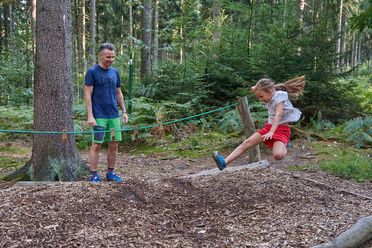 Ein Erwachsener und ein Kind spielen im Wald, eines springt von einem Baumstamm, das andere schaut zu.