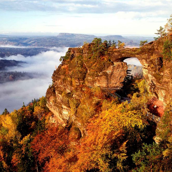 Felsformation mit einem natürlichen Bogen, umgeben von buntem Laub und Nebel in der Ferne.