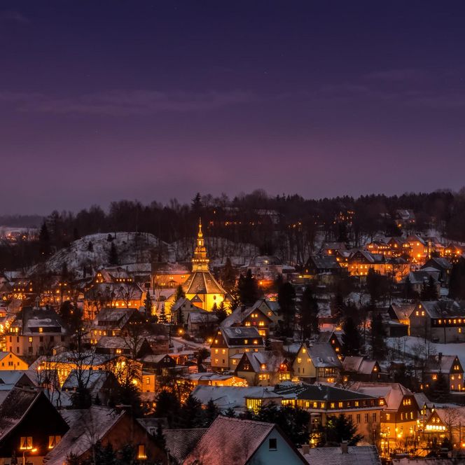 Winterliche Stadtansicht bei Dämmerung, beleuchtete Häuser und schneebedeckte Landschaft.