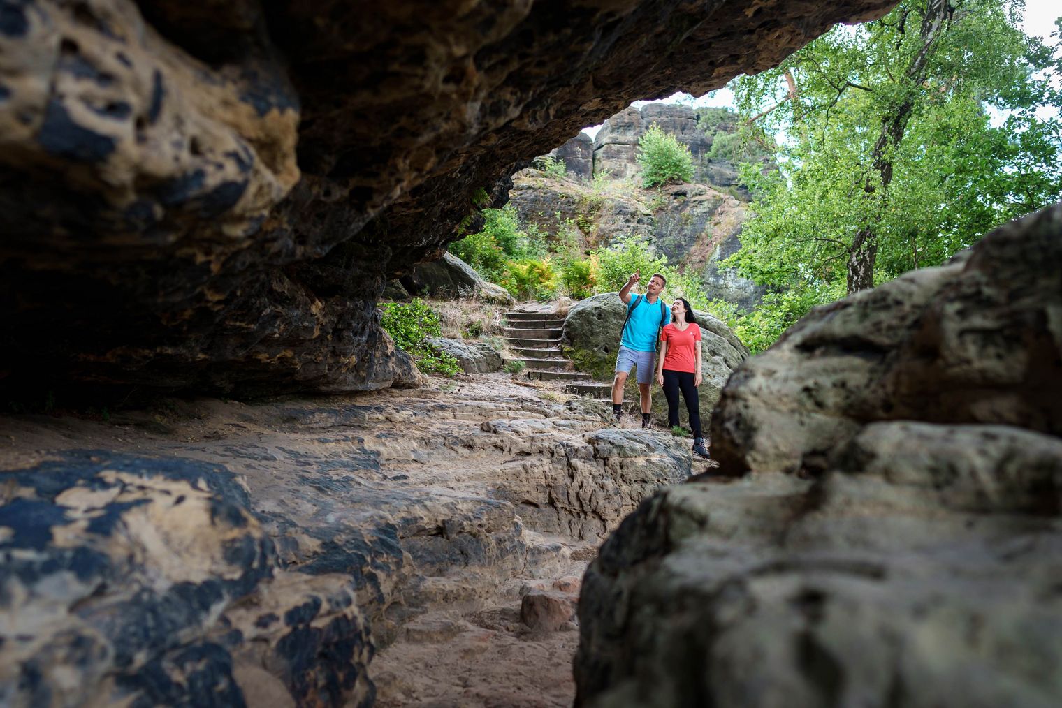 Zwei Personen wandern durch eine felsige Landschaft mit grünen Pflanzen im Hintergrund.