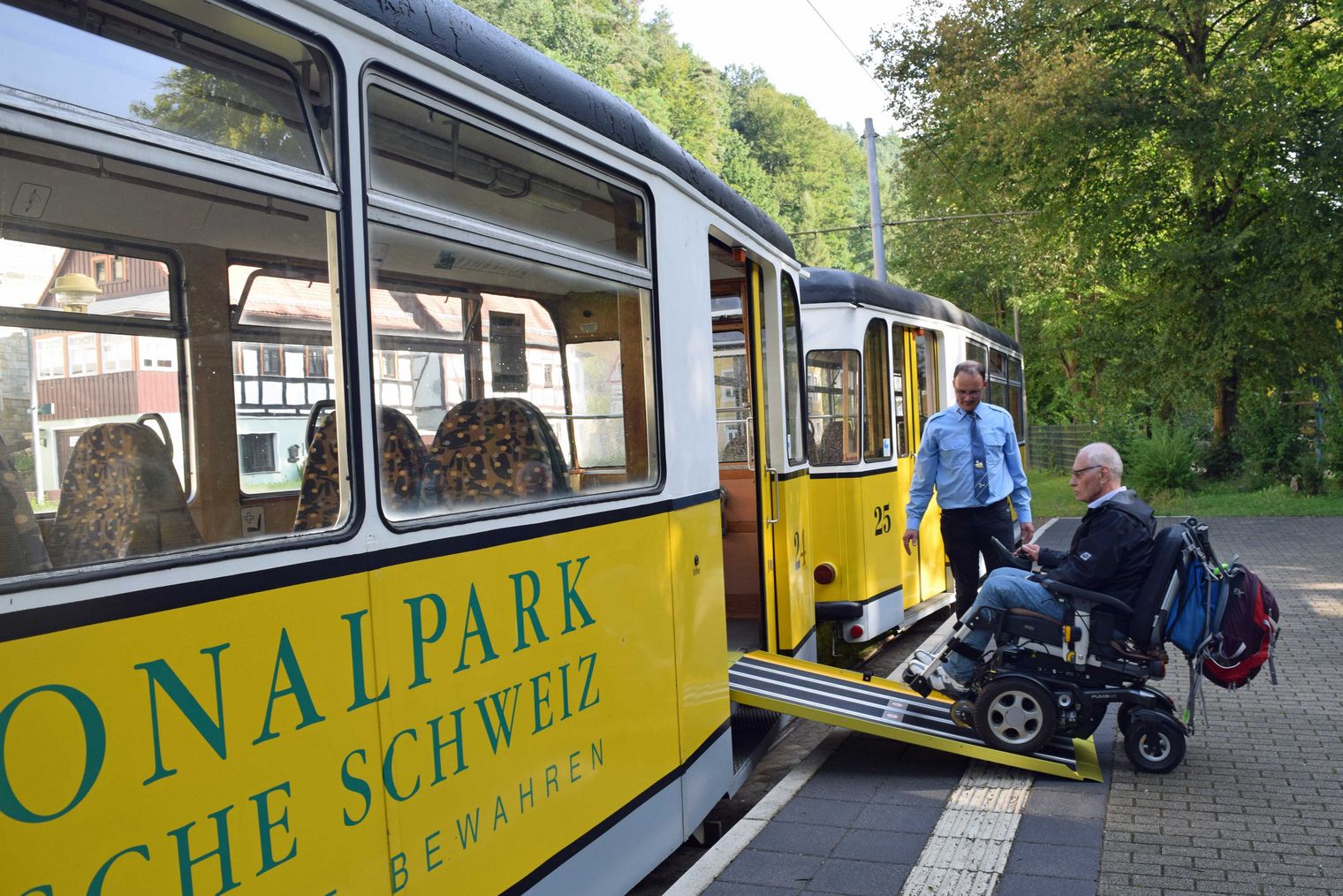 Rollstuhlfahrer fährt mit einer Rampe in eine gelbe Bahn, während ein Mitarbeiter hilft. Grüne Bäume im Hintergrund.