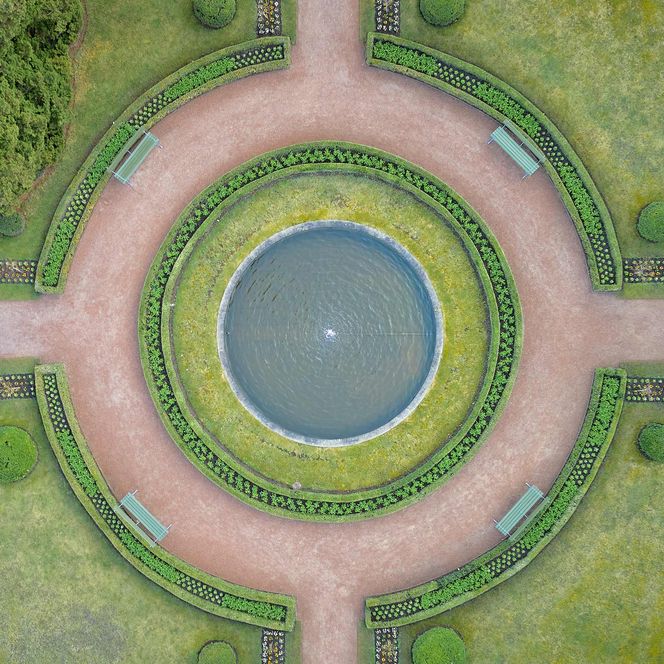 Runder Teich in einem symmetrischen Garten mit Wegen und grünen Hecken.