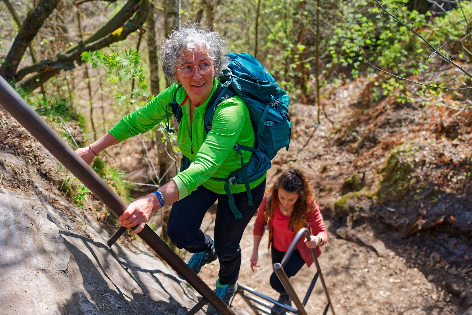 Zwei Wanderer mit Rucksack kraxeln eine steile Treppe in der Natur hinauf, umgeben von Bäumen und Pflanzen.