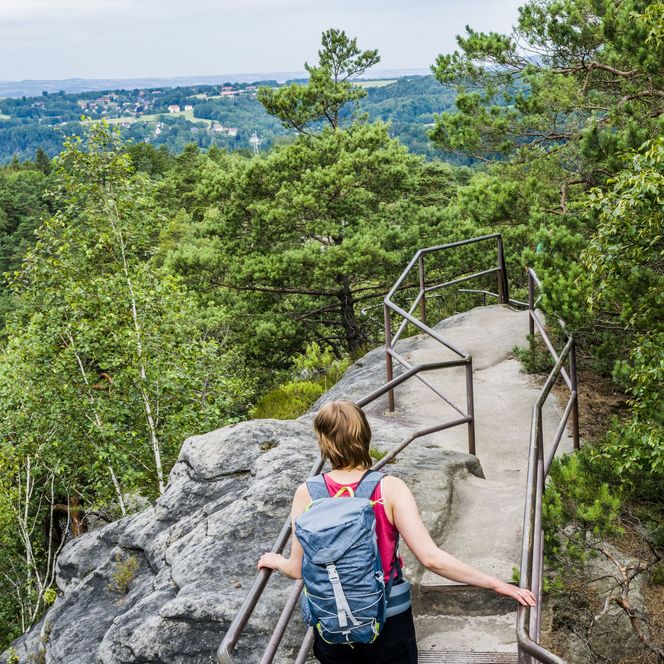 Frau mit Rucksack geht auf einem schmalen Weg zwischen Bäumen und Felsen mit Aussicht ins Tal.