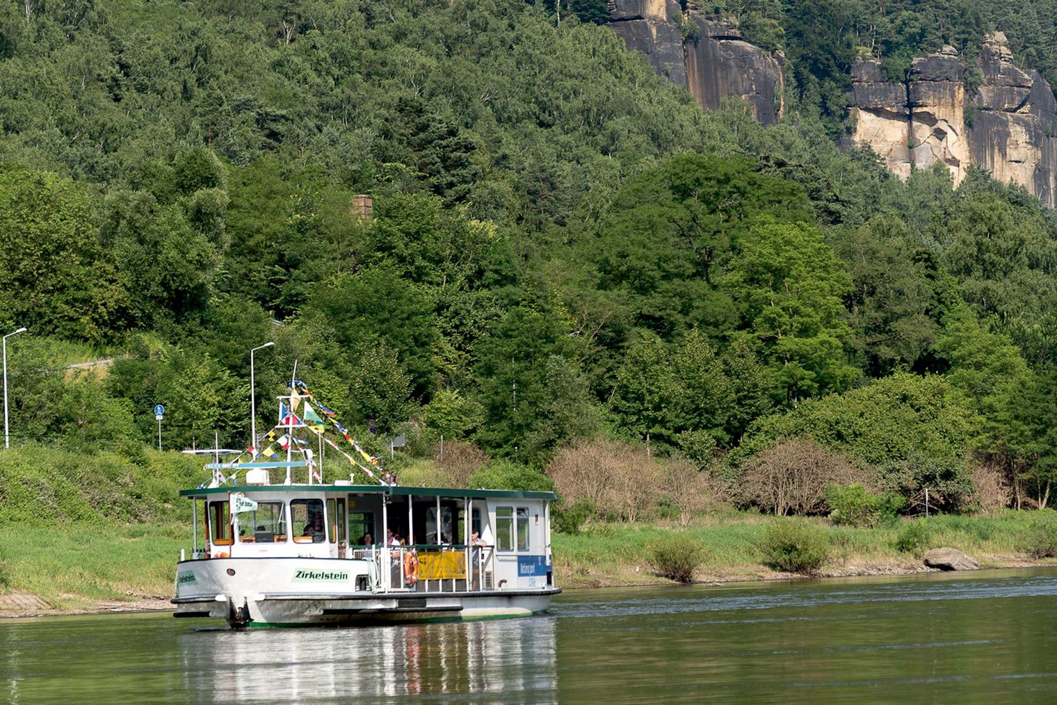 Das Ausflugsschiff "Wanderschiff" fährt auf der ruhigen Elbe, umgeben von grünen Bäumen und steilen Felsen.