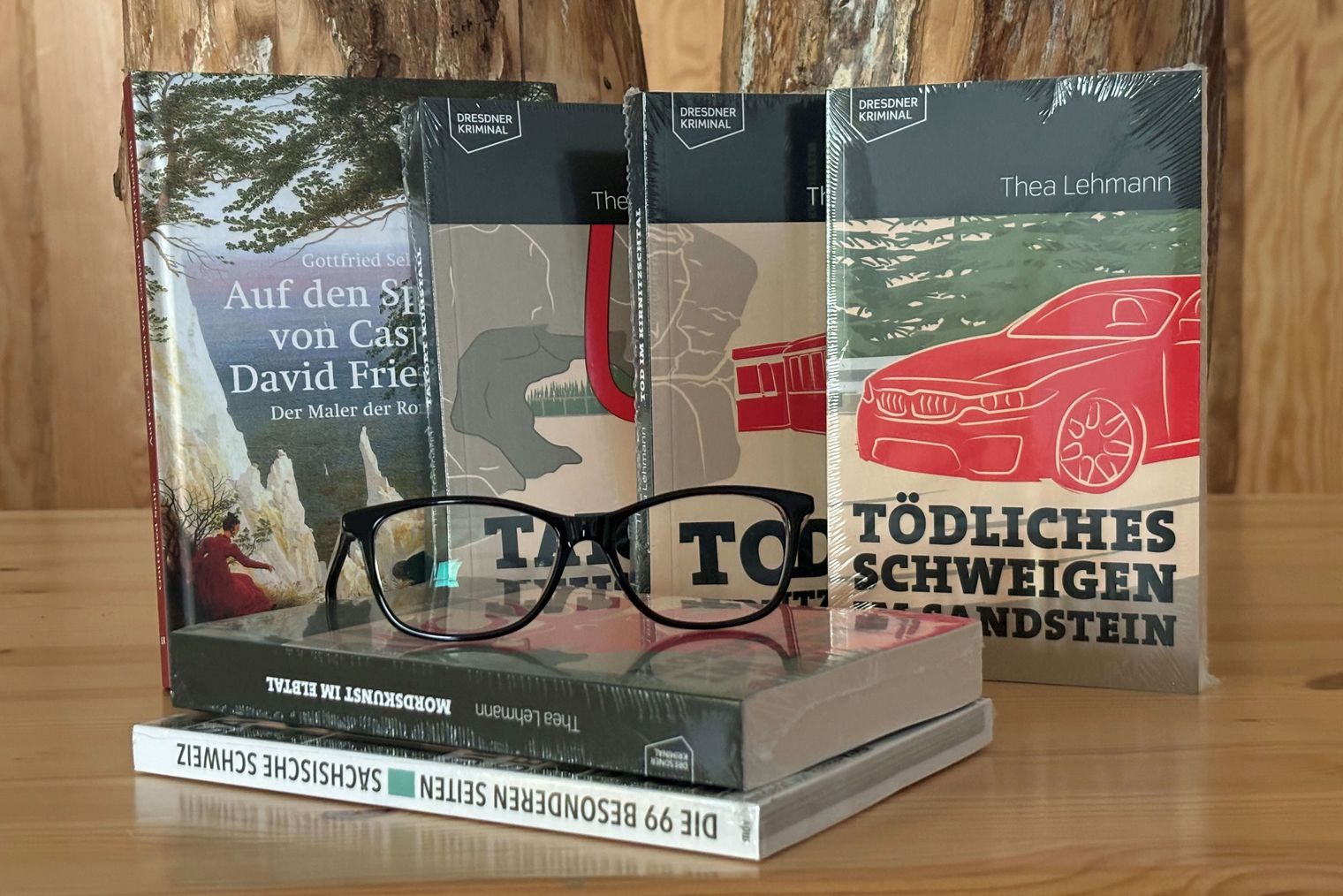 Sechs Bücher stehen auf einem Holztisch, davor liegt eine Lesebrille.