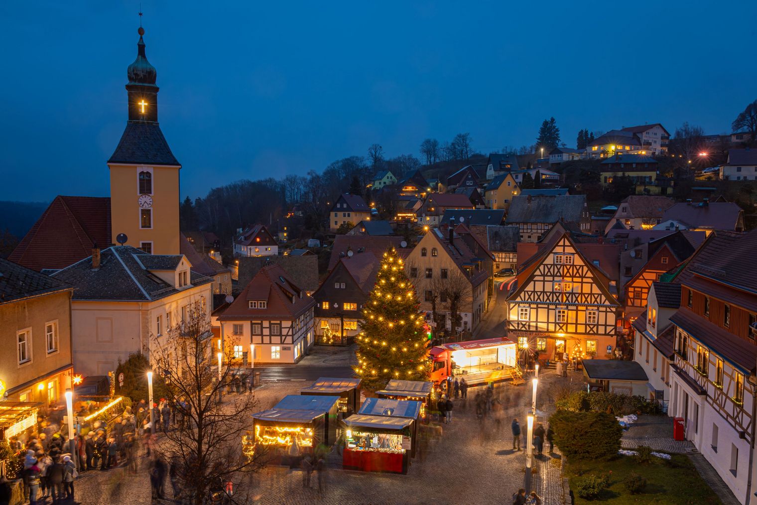 Weihnachtsmarkt mit festlich beleuchteten Ständen und einem großen Weihnachtsbaum in einer Stadt bei Dämmerung.