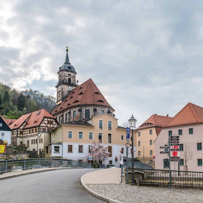 Historische Gebäude mit roten Dächern und einer Kirche, umgeben von bewaldeten Hügeln und bewölktem Himmel.