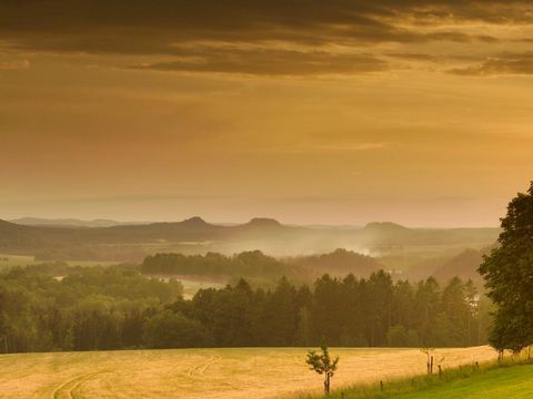 Blick über grünes Tal mit verstreuten Häusern und Hügeln im Hintergrund unter orangenem Himmel.