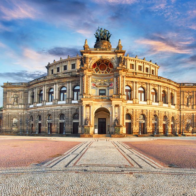 Historisches Gebäude mit aufwendiger Fassade und Uhr, umgeben von einem Platz und blauem Himmel.