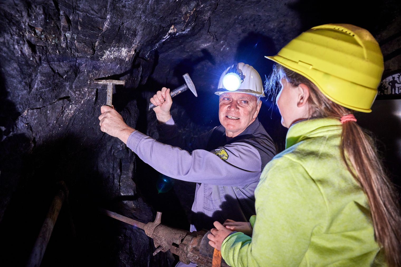 Zwei Personen in einem Bergwerk, eine mit Helm und Stirnlampe, die mit einem Hammer auf einen Felsen schlägt.