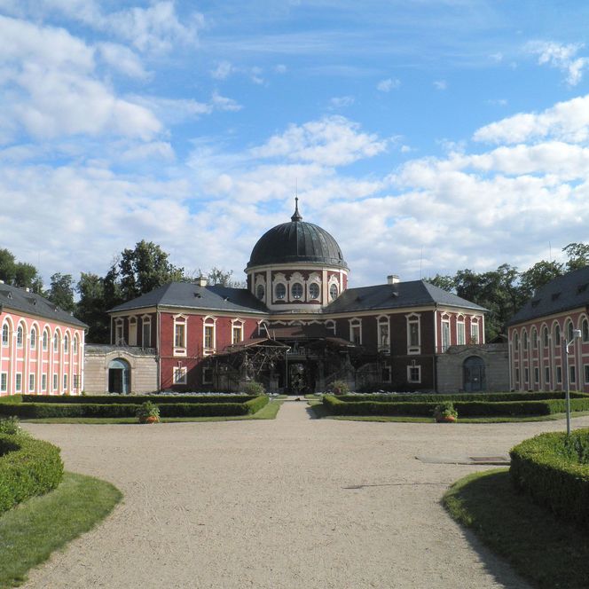 Blick auf ein historisches Gebäude mit Kuppel, umgeben von gepflegten Gärten und blauem Himmel.