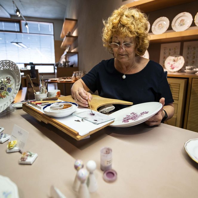 Frau mit lockigem Haar bemalt Geschirr auf einem Tisch, umgeben von Porzellanfiguren und Geschirr.
