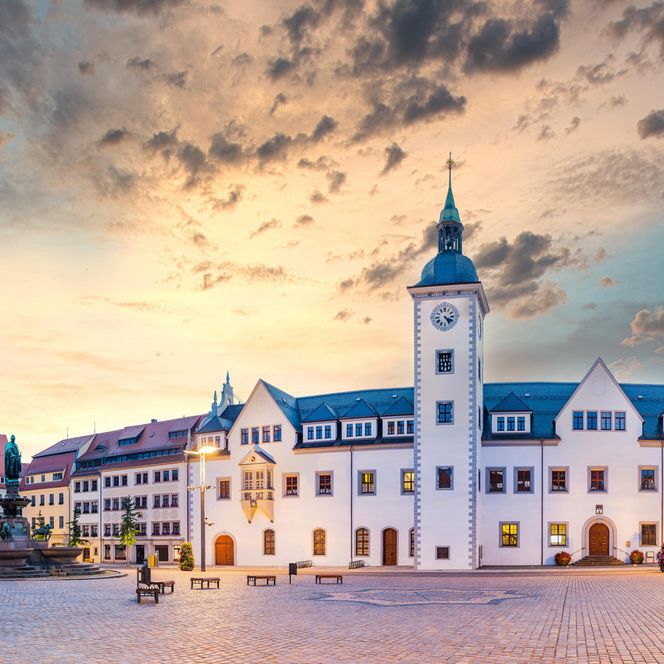 Historisches Gebäude mit Turm und blauer Dachkonstruktion, umgeben von Platz und Wolkenhimmel.