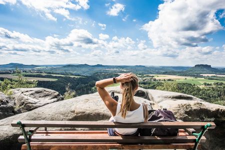Frau mit langen Haaren sitzt auf einer Bank und blickt auf eine weite Landschaft mit Hügeln und Wolken.