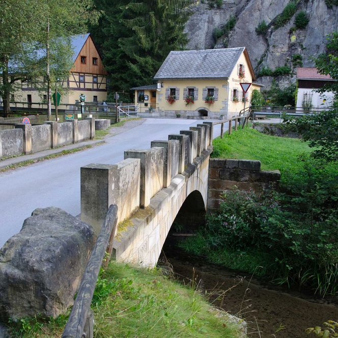 Steinbrücke über einen kleinen Fluss, umgeben von Bäumen und historischen Gebäuden.