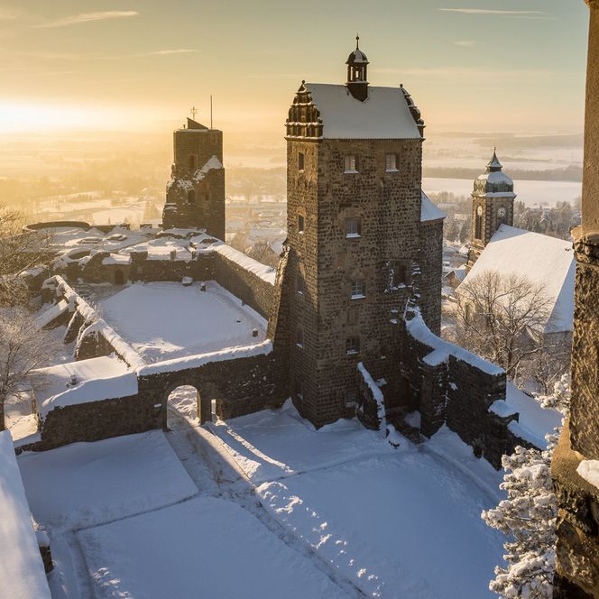 Schneebedeckte Burgruine mit Türmen und Sonnenaufgang im Hintergrund.