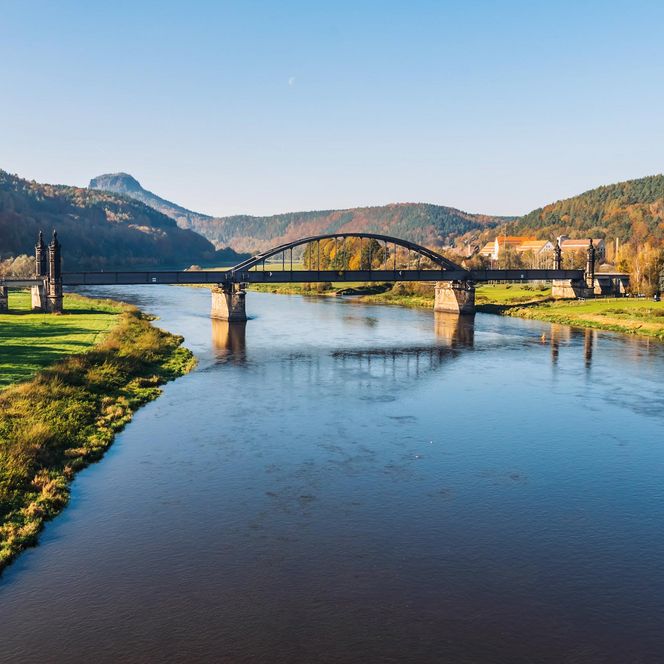 Brücke über einen ruhigen Fluss, umgeben von grünen Wiesen und Hügeln im Hintergrund.