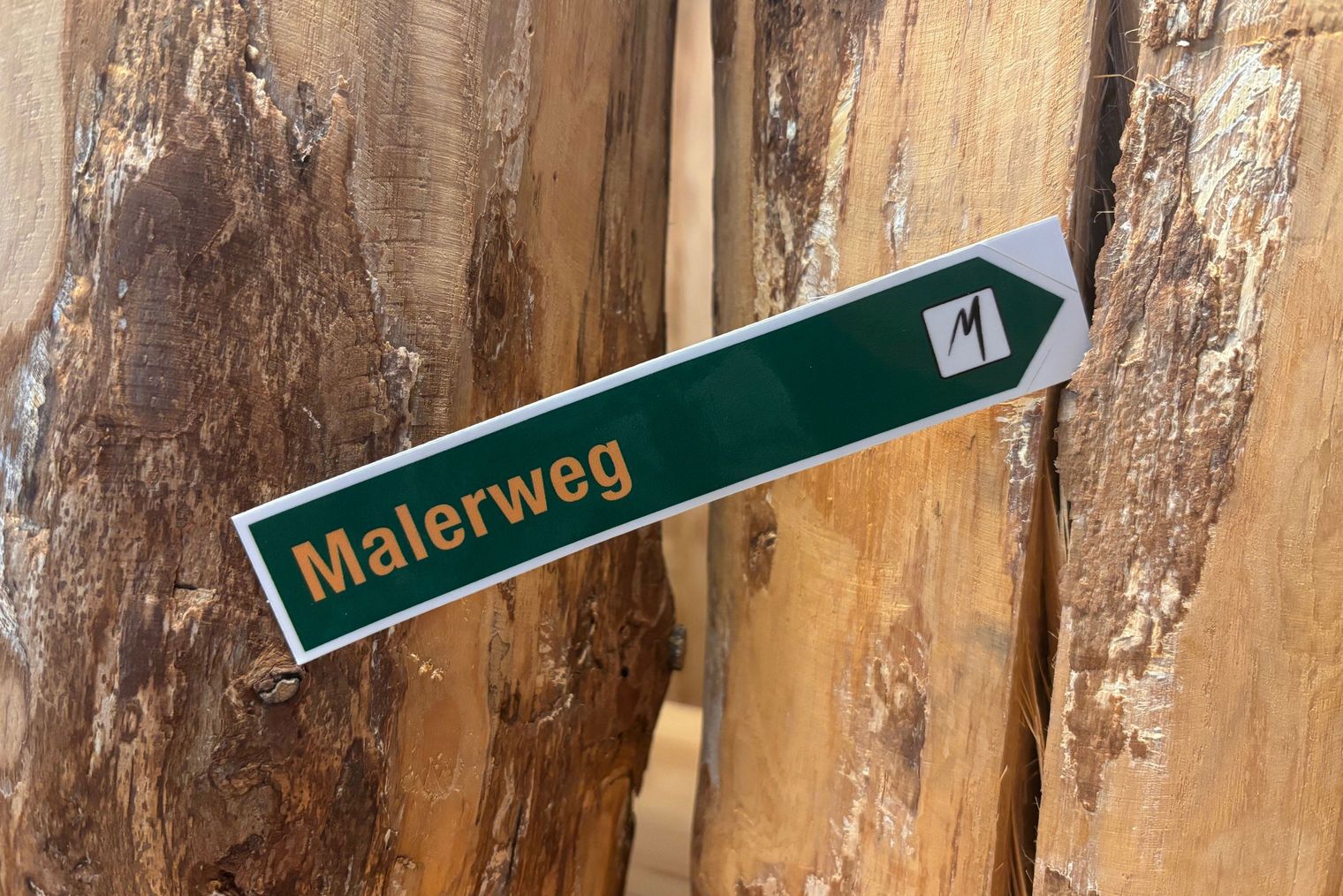 Wegweiser mit der Aufschrift "Malerweg", im Hintergrund zwei Baumstämmen.