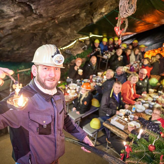 Mann mit Stirnlampe hält eine Fackel, umgeben von Menschen an einem festlich dekorierten Tisch in einer Höhle.
