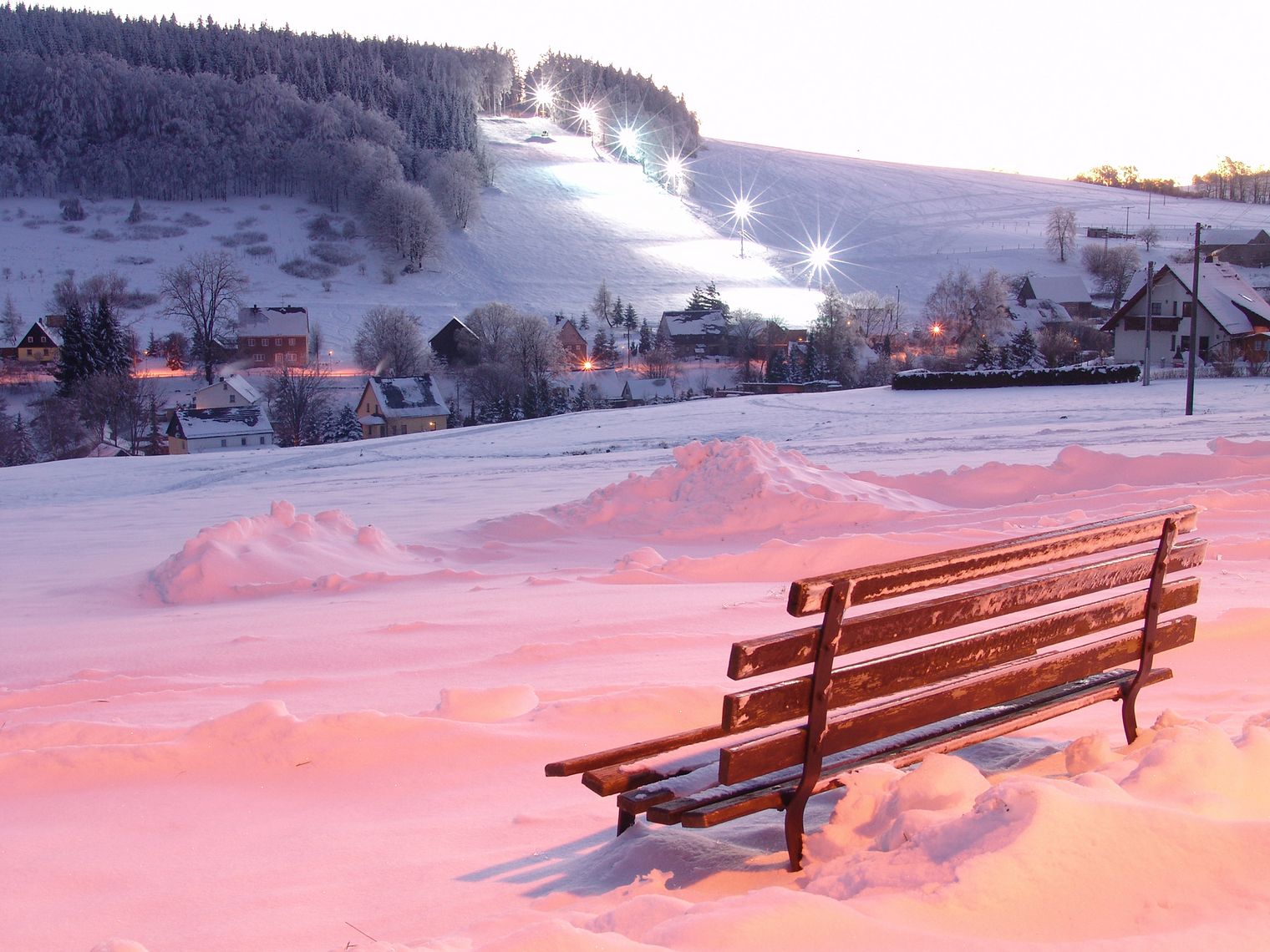 Schneebedeckte Landschaft mit einer Holzbank im Vordergrund und schneebedeckten Hügeln im Hintergrund. Skihang in Rugiswalde