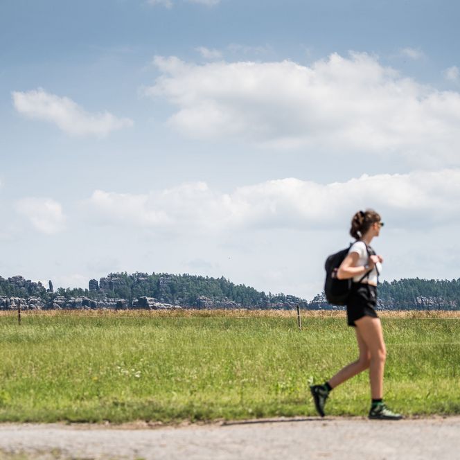 Junge Wanderin mit Rucksack geht auf einem Weg durch eine grüne Landschaft unter blauem Himmel.