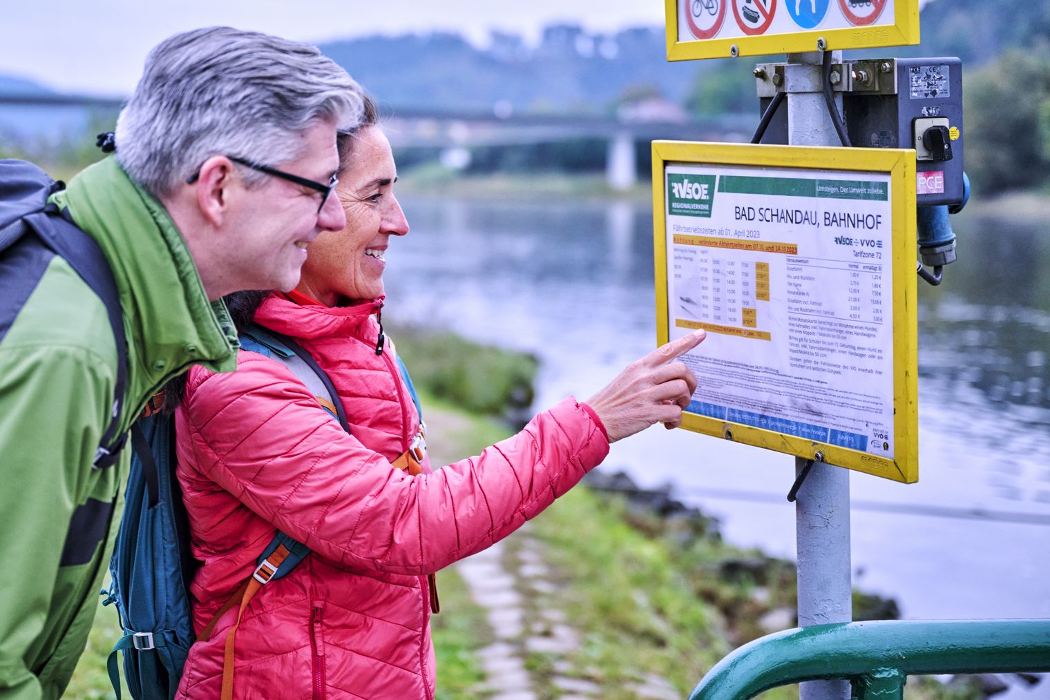 Frau in roter Jacke liest Wanderkarte am Fähranleger Nationalparkbahnhof Bad Schandau, Fluss im Hintergrund.
