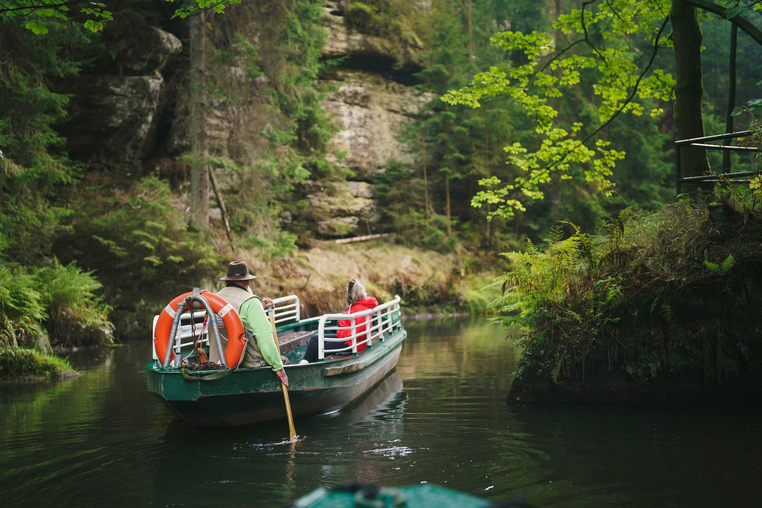 Ein Kahnfahrer paddelt Touristen in einem Boot der Kahnfahrt Obere Schleuse durch eine grüne, bewaldete Schlucht mit Felsen und ruhigem Wasser.