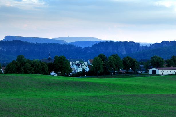 Gruene Wiesen im Vordergrund, bewaldete Berge im Hintergrund unter bewölktem Himmel.
