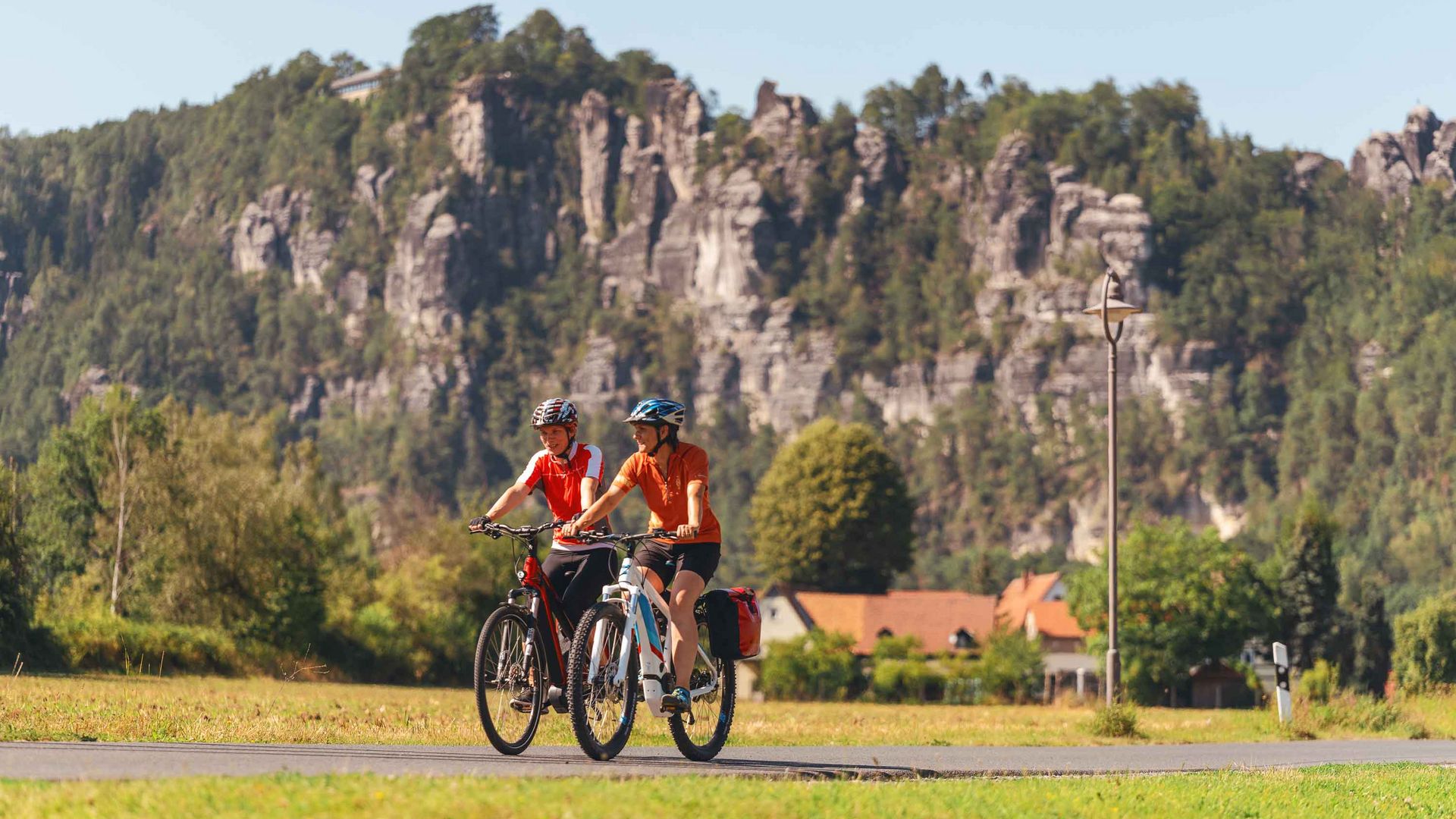 Zwei Radfahrerinnen in roten Shirts fahren auf einem Weg, im Hintergrund Felsen und Bäume.