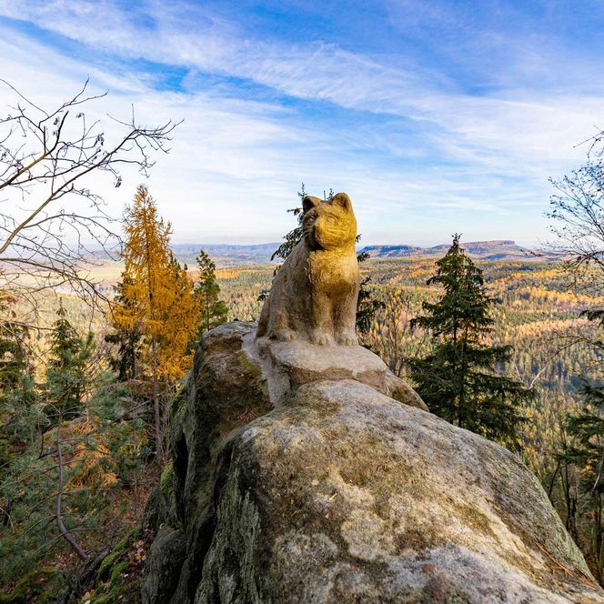 Aus Sandstein geformte Statue einer Katze sitzt auf einem Felsen mit Blick auf eine bewaldete Landschaft und einen blauen Himmel.