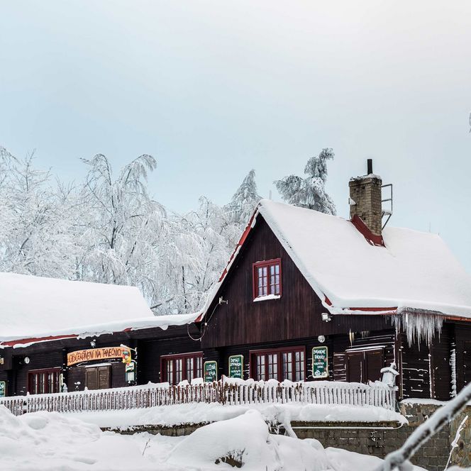 Holzhaus im Schnee, umgeben von schneebedeckten Bäumen und einem klaren Himmel.