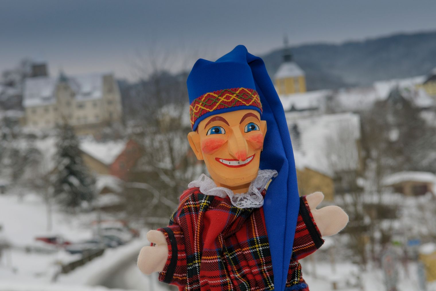 Puppe mit blauer Mütze und rotem Kleid vor einer verschneiten Stadtlandschaft.