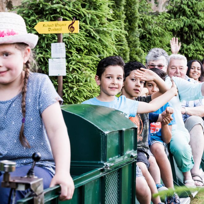Kinder sitzen auf einer kleinen Eisenbahn, lächeln und haben Spaß, umgeben von grünen Pflanzen.