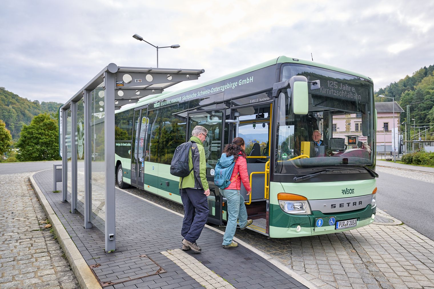 Zwei Personen steigen in einen grünen Bus an einer Haltestelle ein.