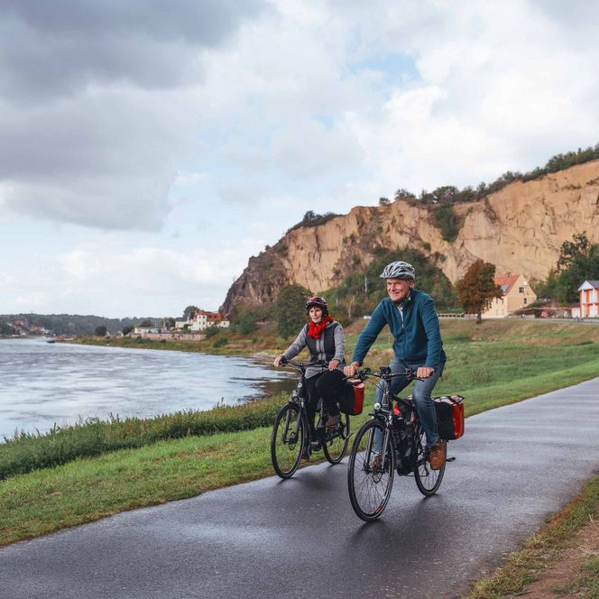 Zwei Radfahrer fahren entlang eines Flusses, umgeben von einer steilen Klippe und Bäumen.