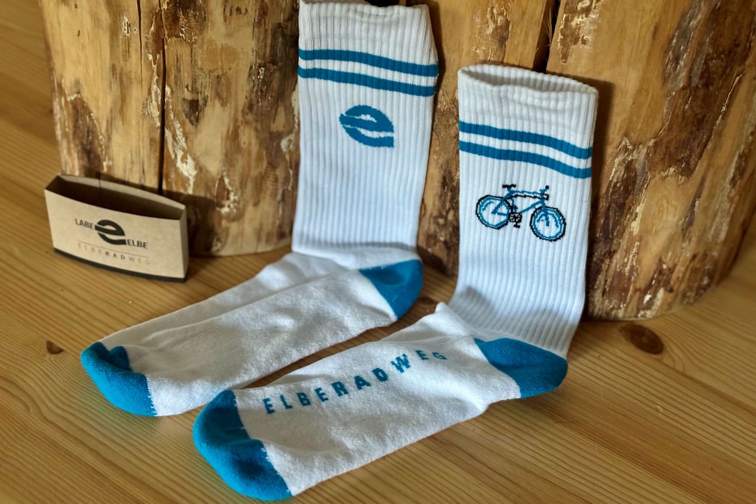Auf einem Holztisch liegen weiße Socken mit blauem Logo und Fahrradmotiv, neben einer kleinen Verpackung.
