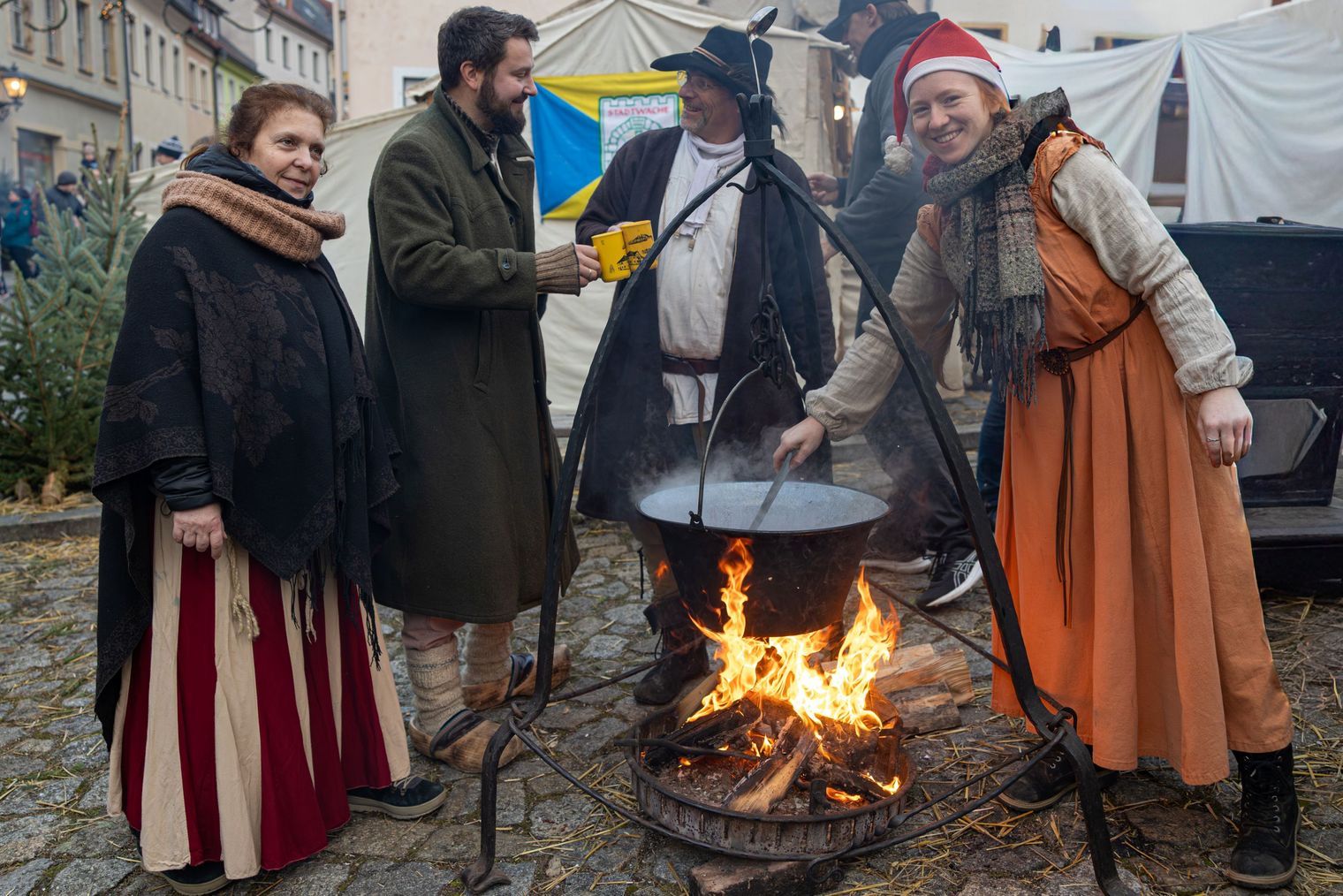 Vier Personen in historischen Kostümen stehen um einen Kessel über einem Feuer.