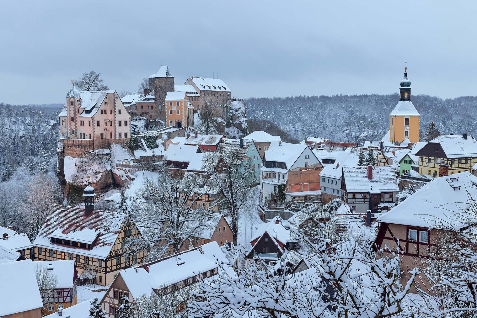 Schneebedeckte Häuser und eine Kirche in einem winterlichen Dorf, umgeben von verschneiten Bäumen.