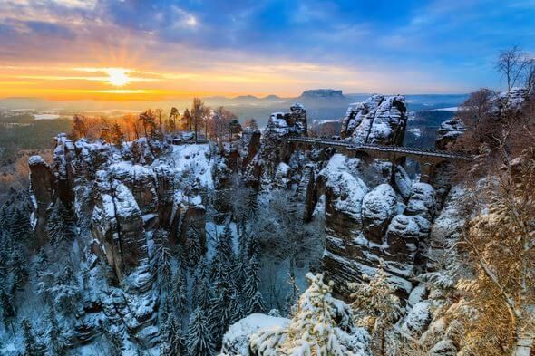 Winterlandschaft mit schneebedeckter Bastei-Brücke, Felsen und Bäumen im Morgenlicht.