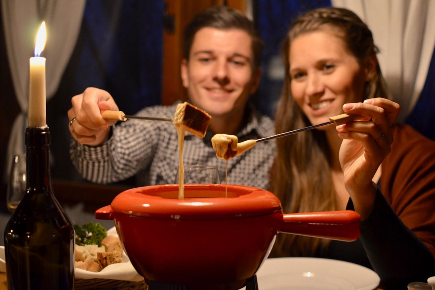 Paar sitzt am Tisch, genießt Fondue mit einer Gabel, Kerze und Weinflasche im Hintergrund.