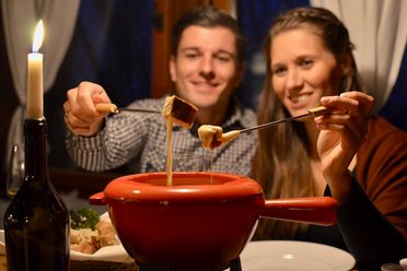 Paar sitzt am Tisch, genießt Fondue mit einer Gabel, Kerze und Weinflasche im Hintergrund.
