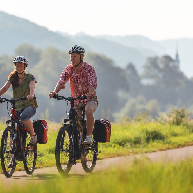 Zwei Radfahrer auf einem Weg durch eine grüne Landschaft, umgeben von Hügeln.
