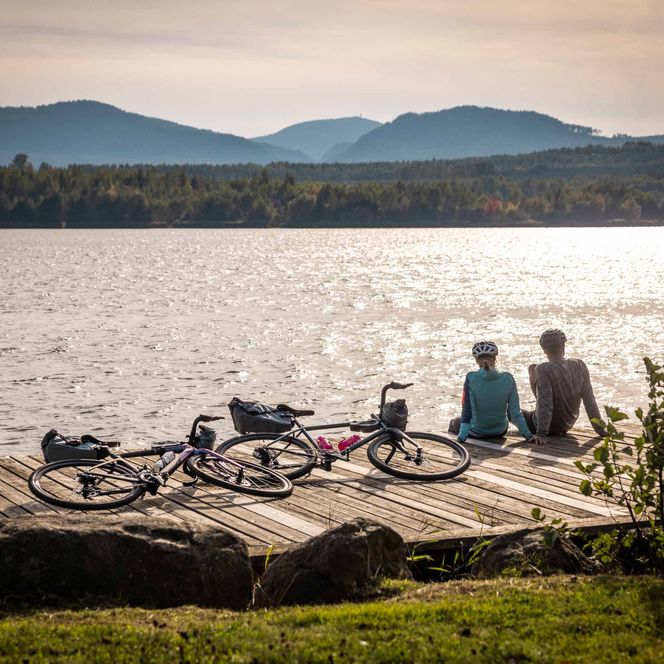 Zwei Personen sitzen auf einem Steg am Olbersdorfer See, Fahrräder liegen daneben, Berge im Hintergrund.