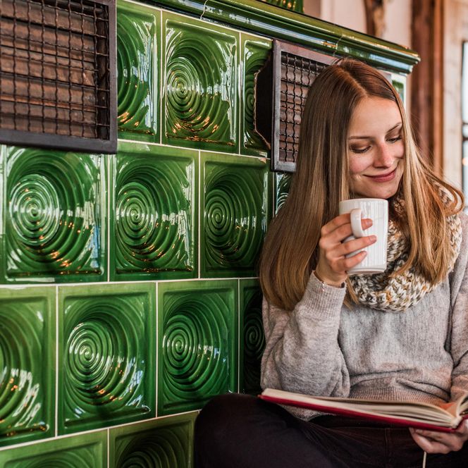 Frau mit langen Haaren sitzt an einer grünen Kachelwand und liest ein Buch, während sie eine Tasse hält.